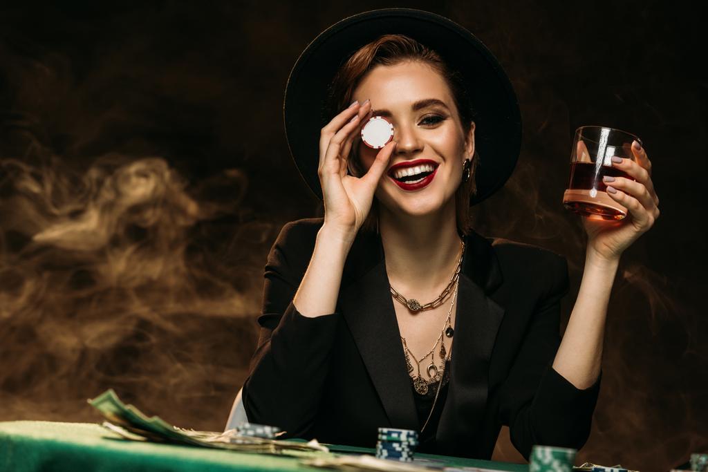 glückliches attraktives Mädchen in Jacke und Hut, das ein Glas Whiskey am Pokertisch hält und das Auge mit einem Casino-Chip bedeckt - Foto, Bild