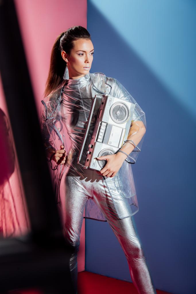 fashionable young woman in metallic bodysuit and raincoat posing with retro boombox on pink and blue background - Photo, Image