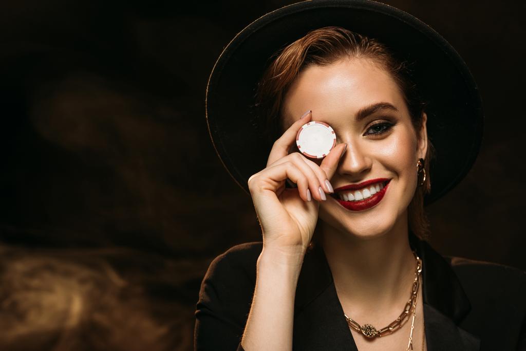 ευτυχισμένος ελκυστικό κορίτσι στο σακάκι και καπέλο που καλύπτει το μάτι με το τσιπ πόκερ και να βλέπουν τα φωτογραφικών μηχανών - Φωτογραφία, εικόνα