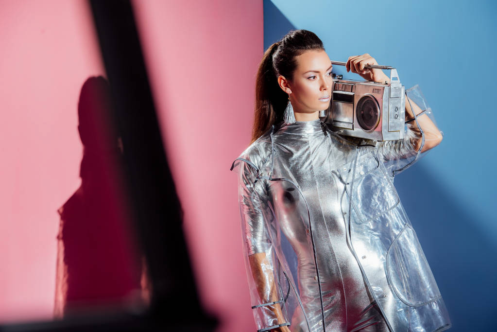 stylish woman in metallic bodysuit and raincoat posing with retro boombox on pink and blue background - Photo, Image