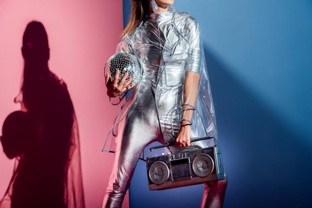 bijgesneden weergave van modieuze vrouw in zilveren Romper en regenjas poseren met boombox en disco bal op roze en blauwe achtergrond - Foto, afbeelding