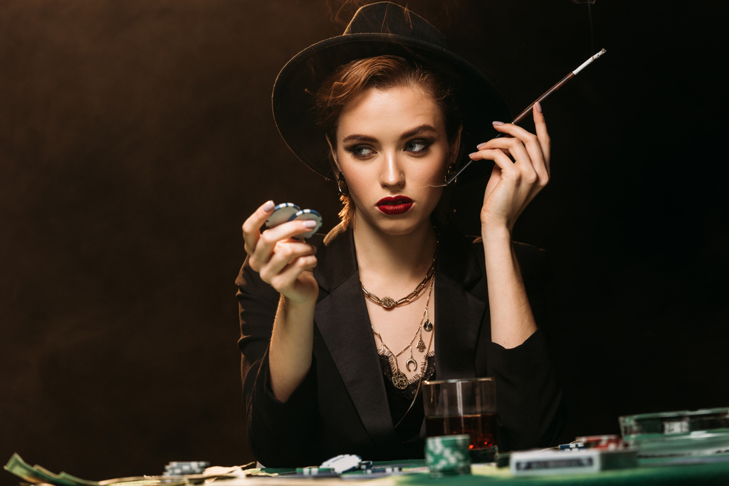 attraktives Mädchen in Jacke und Hut raucht Zigarette am Pokertisch im Casino und schaut weg - Foto, Bild