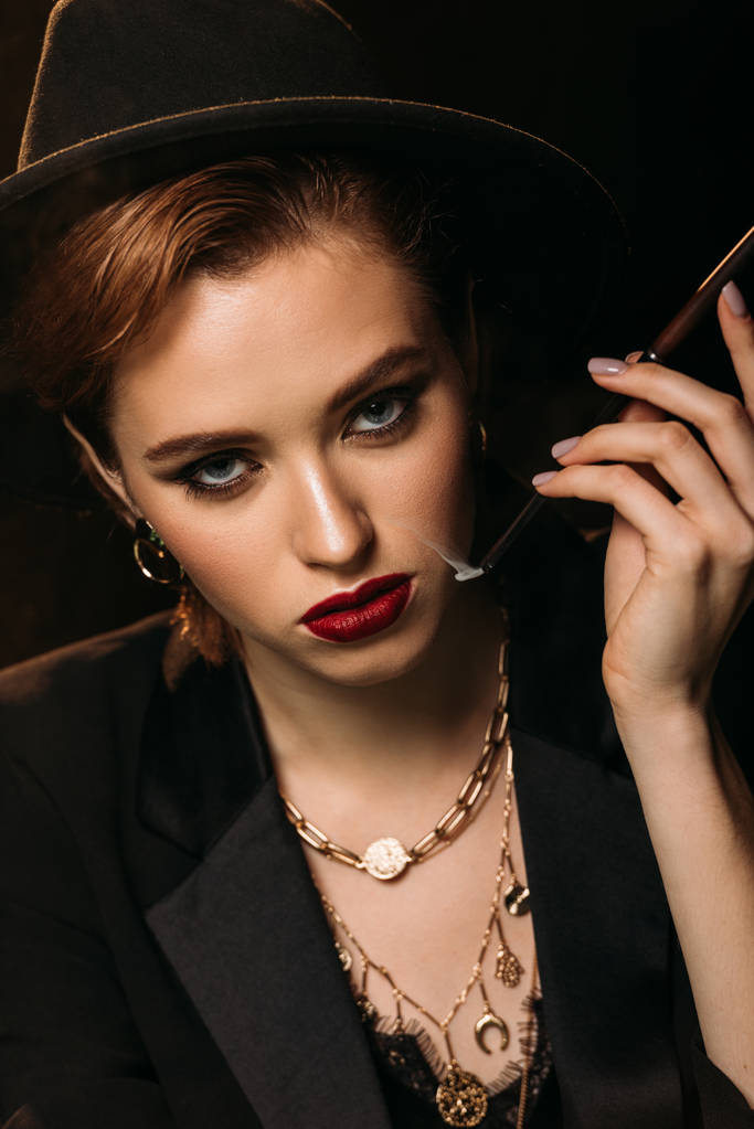 portrait de fille attrayante en veste et chapeau fumer cigarette sur noir et en regardant la caméra
 - Photo, image