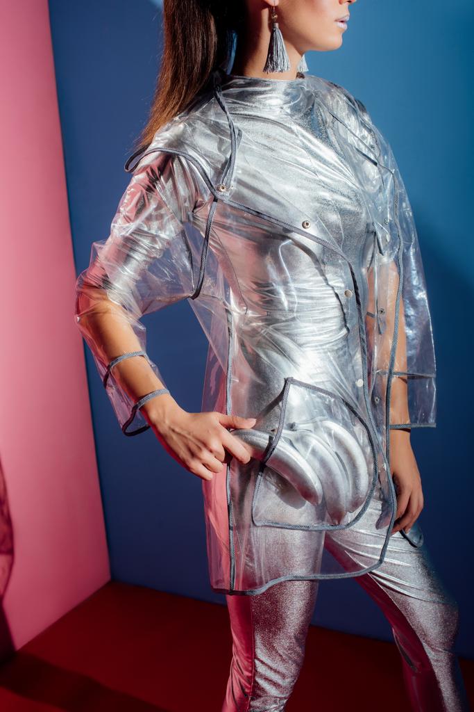 cropped view of girl in metallic bodysuit and raincoat posing with silver bananas for fashion shoot on pink and blue background - Photo, Image
