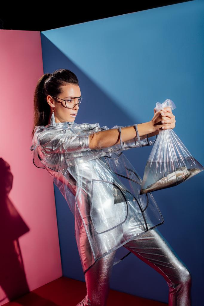 trendy girl in silver bodysuit and raincoat holding plastic bag with fish on pink and blue background - Photo, Image