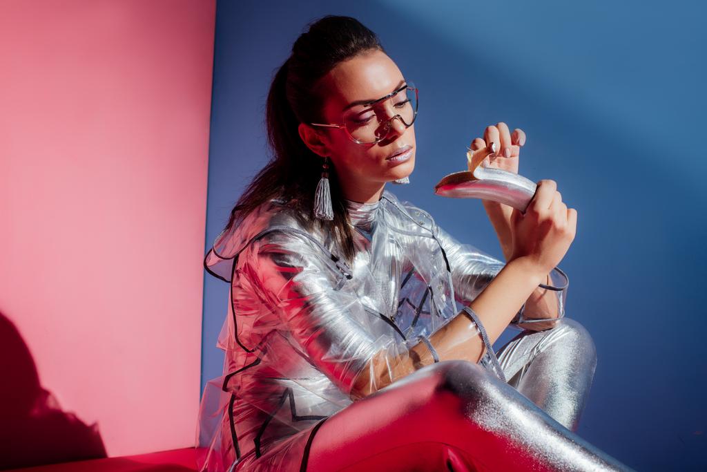fashionable model in metallic bodysuit and raincoat posing with silver banana on pink and blue background - Photo, Image