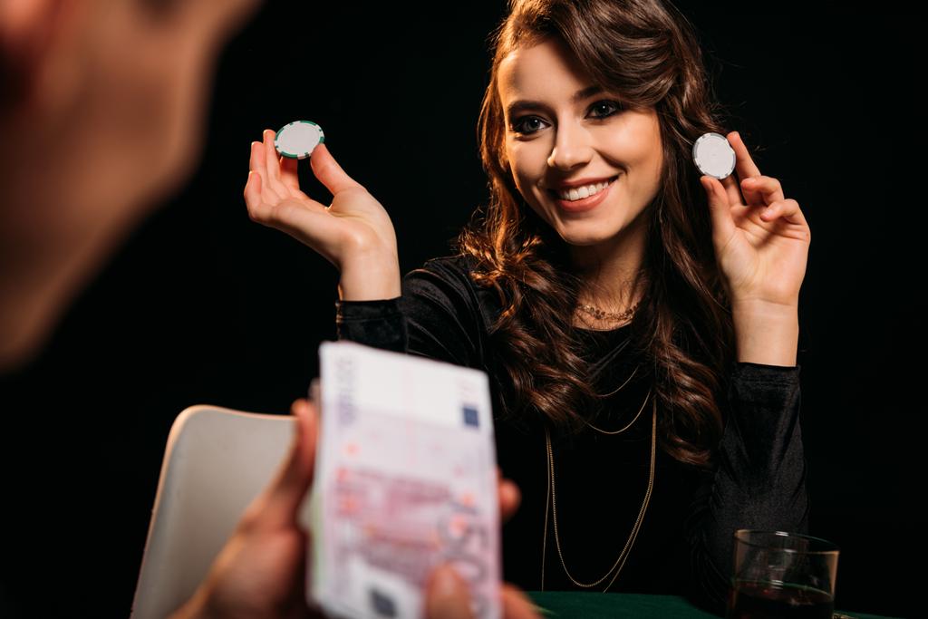 привлекательная улыбающаяся девушка держит фишки для покера и смотрит на мужчину за покерным столом в казино
 - Фото, изображение