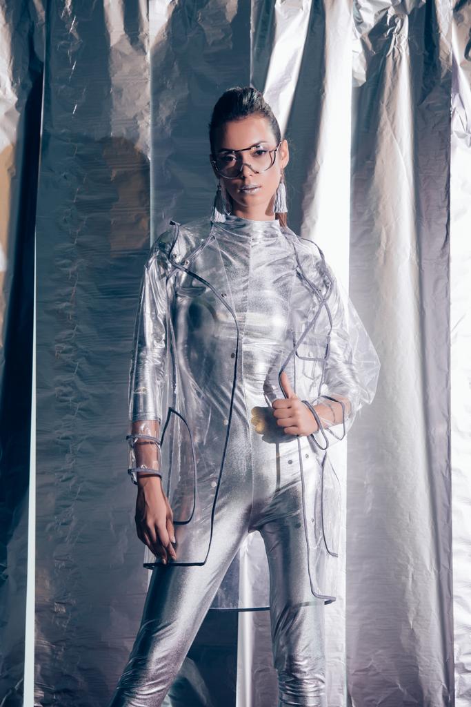 modelo jovem atraente posando em bodysuit prata e capa de chuva em fundo metálico
 - Foto, Imagem