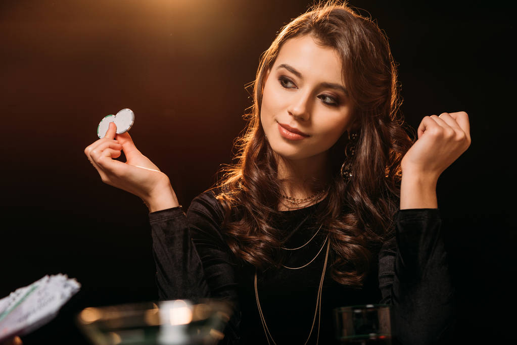 приваблива дівчина тримає покерні чіпи і дивиться в сторону покерного столу в казино
 - Фото, зображення