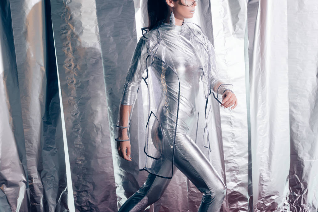 abgeschnittene Ansicht einer stilvollen Frau, die in silbernem Body und Regenmantel auf metallischem Hintergrund posiert - Foto, Bild