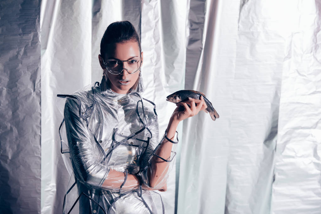 modelo joven atractivo en traje de cuerpo de plata y impermeable sosteniendo peces sobre fondo metálico
 - Foto, Imagen