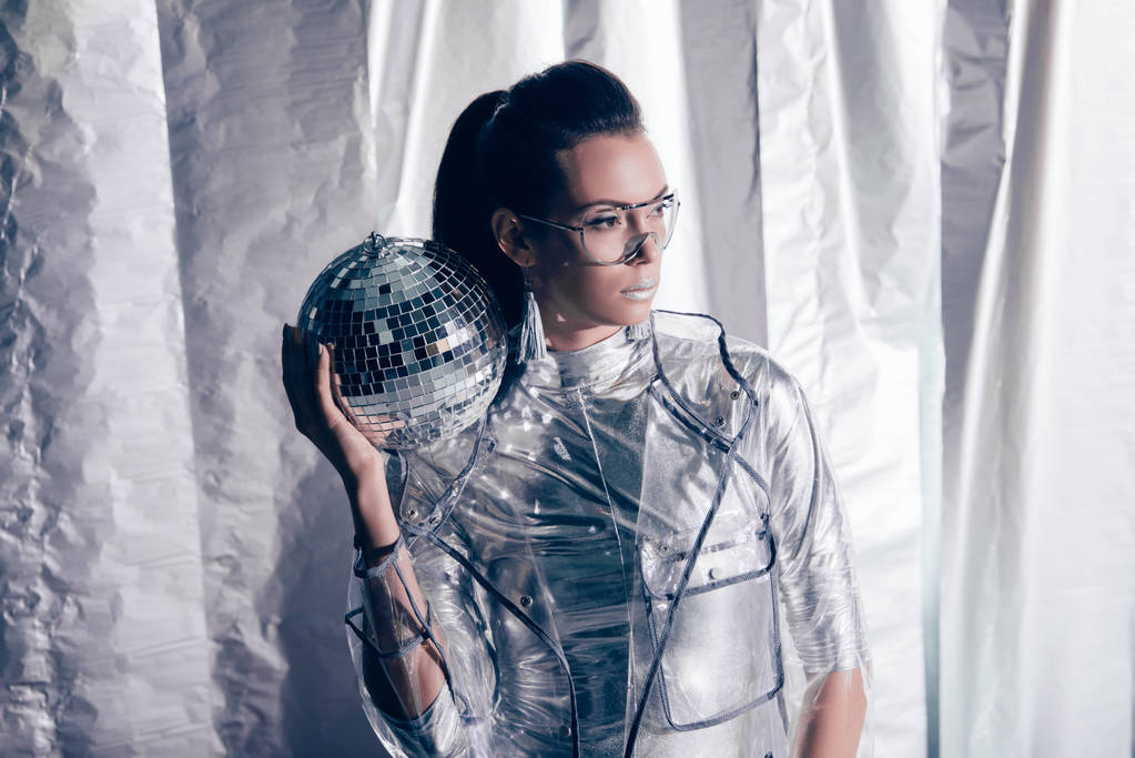 fashionable young woman in silver bodysuit and raincoat posing with disco ball on metallic background - Photo, Image