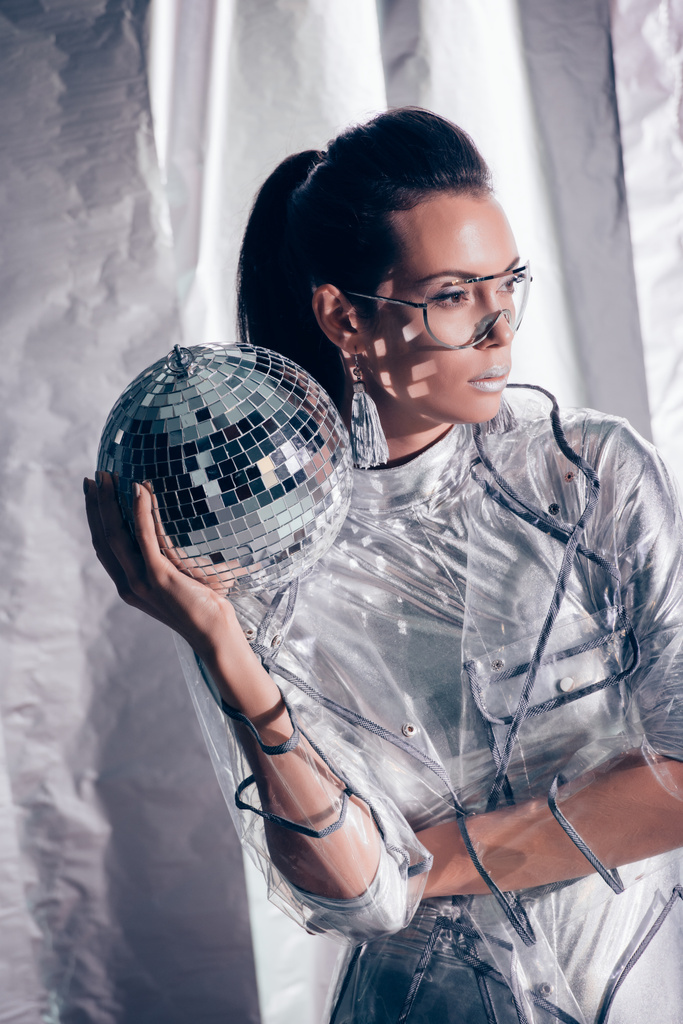 beautiful stylish young woman in silver bodysuit and raincoat posing with disco ball on metallic background - Photo, Image