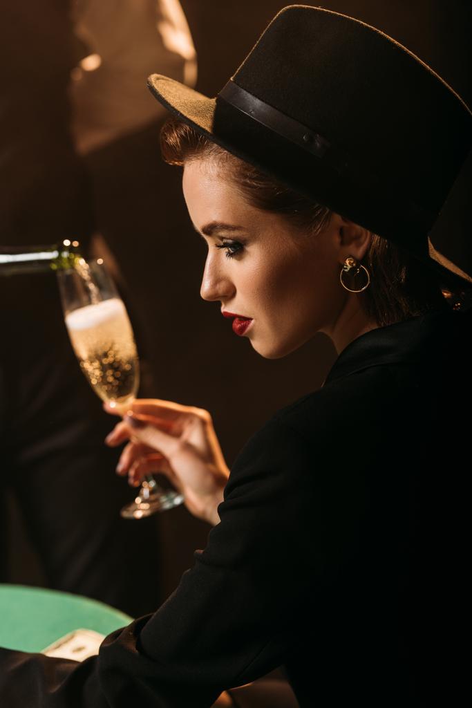 Seitenansicht des Kellners, der Champagner in das Glas eines attraktiven Mädchens in Jacke und Hut im Casino einschenkt - Foto, Bild