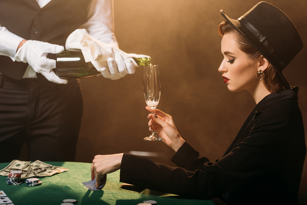 σερβιτόρος έκχυση της σαμπάνιας σε ποτήρι ελκυστικό κορίτσι στο σακάκι και καπέλο ενώ παίζει πόκερ στο καζίνο - Φωτογραφία, εικόνα