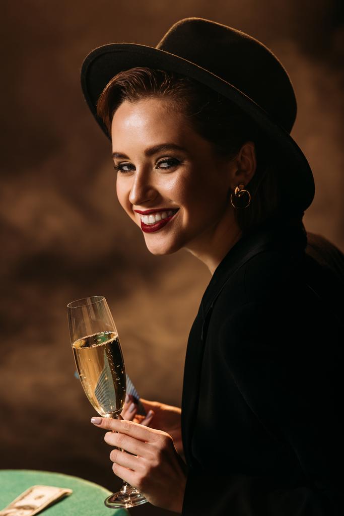 glimlachend aantrekkelijk meisje in jas en hoed bedrijf glas champagne aan pokertafel in het casino en de camera kijken - Foto, afbeelding
