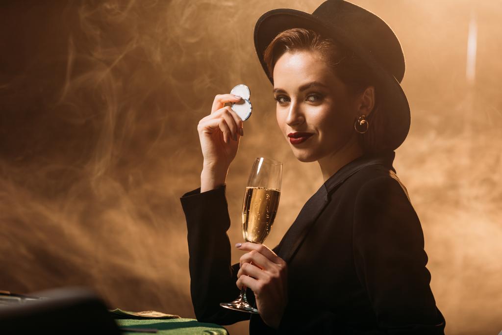 attraente ragazza in giacca e cappello in possesso di un bicchiere di champagne e poker chips a tavola nel casinò
 - Foto, immagini