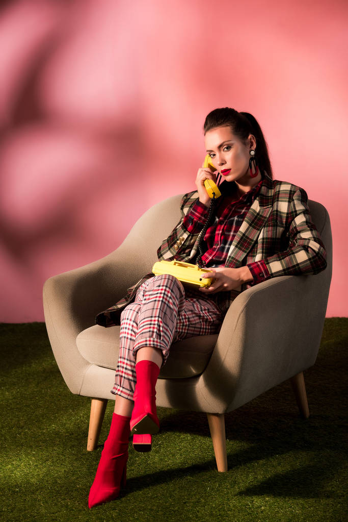 modische Frau im karierten Anzug posiert im Sessel mit Retro-Telefon auf rosa Hintergrund - Foto, Bild