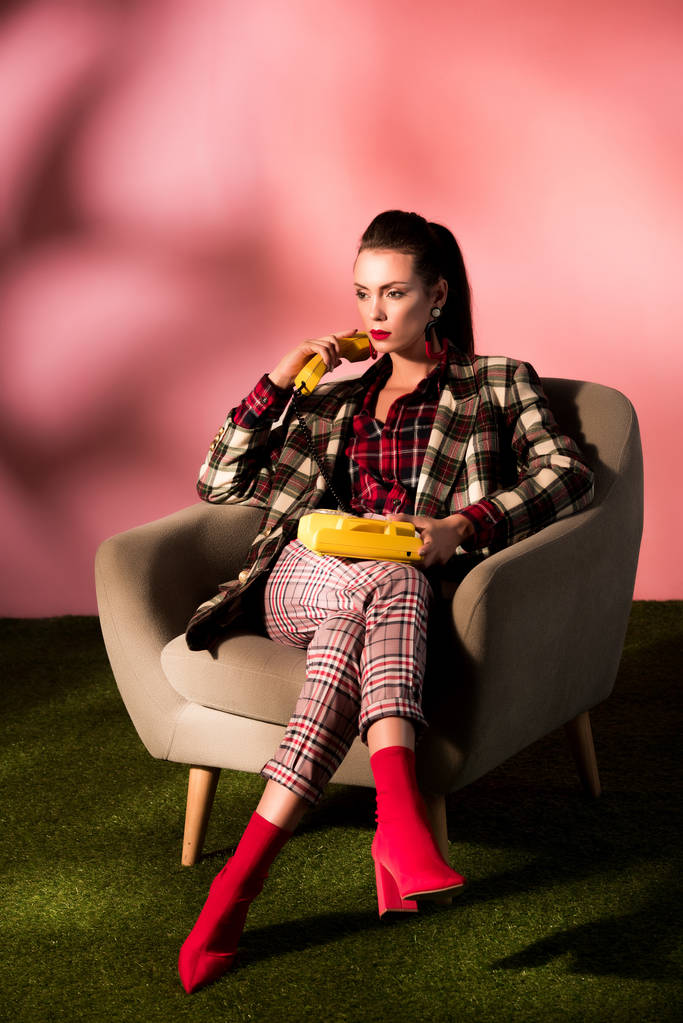 attractive woman in checkered suit posing in armchair with rotary phone on pink background - Photo, Image