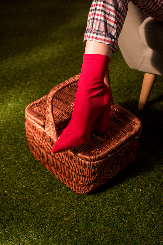 gedeeltelijke weergave van modieuze vrouw in rode schoen poseren met mand op het groene tapijt - Foto, afbeelding