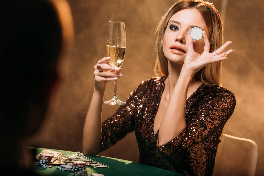 привлекательная девушка держит бокал шампанского и закрывает глаза фишкой для покера за столом в казино
 - Фото, изображение