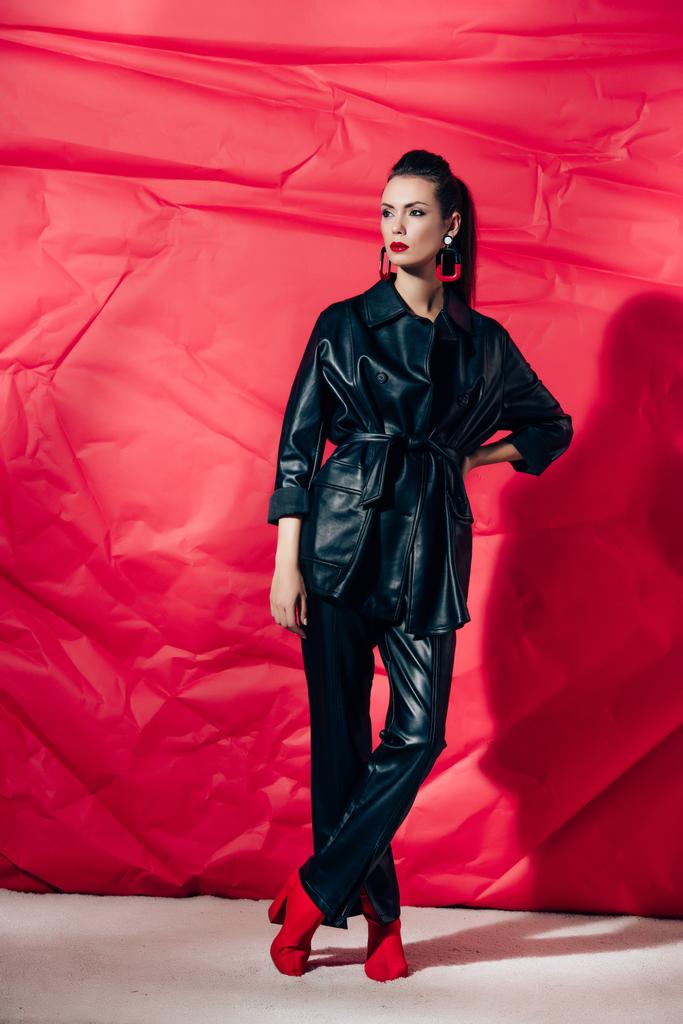 schöne junge Frau posiert in schwarzem Lederanzug auf rotem Hintergrund - Foto, Bild