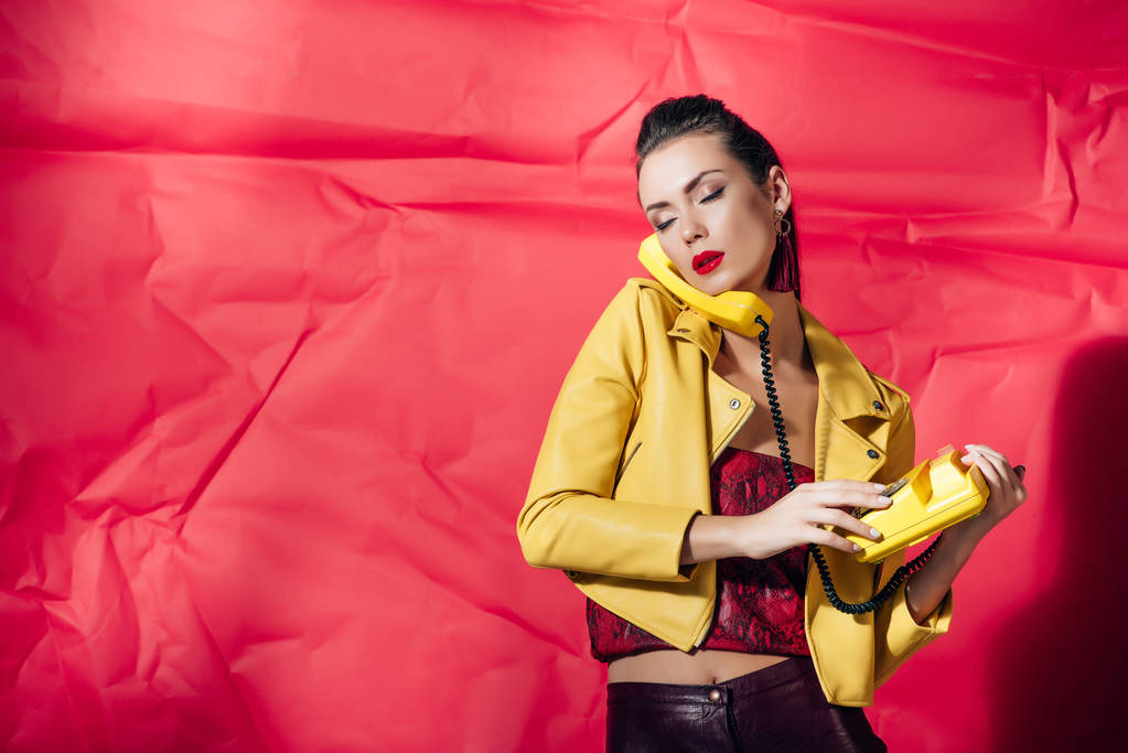beautiful woman in leather jacket posing with yellow rotary phone on red background - Photo, Image