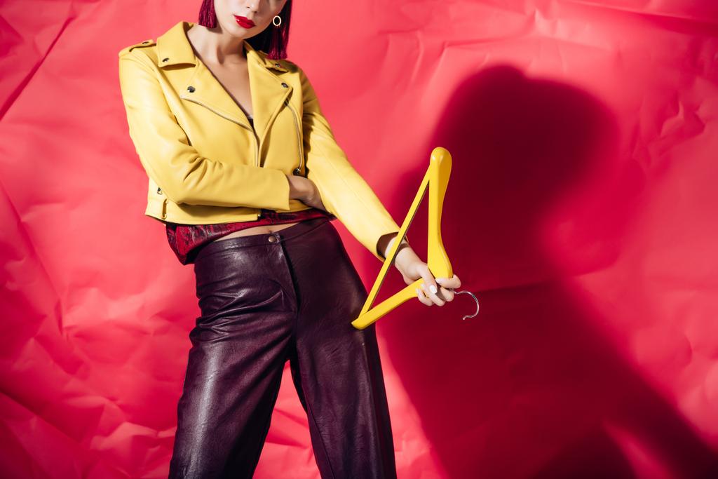 cropped view of model in yellow leather jacket posing with hanger on red background - Photo, Image