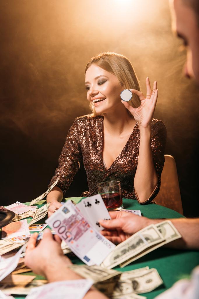 счастливая привлекательная девушка играет в покер с каруселью и держит покерную фишку в казино
 - Фото, изображение