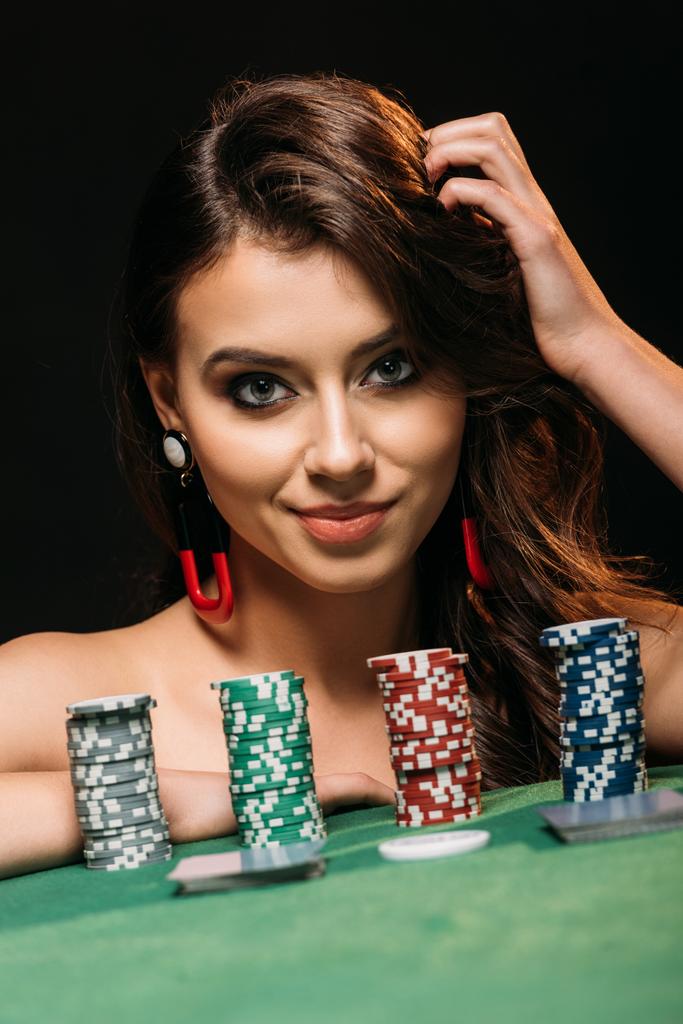 atractiva chica apoyada en la mesa con fichas de póquer y mirando a la cámara aislada en negro
 - Foto, Imagen