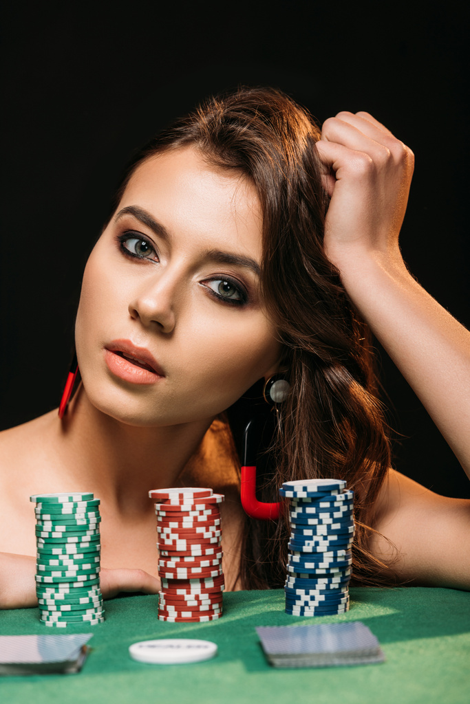 mooi bruin haired meisje leunend op tafel met poker chips en camera geïsoleerd op zwart te kijken - Foto, afbeelding