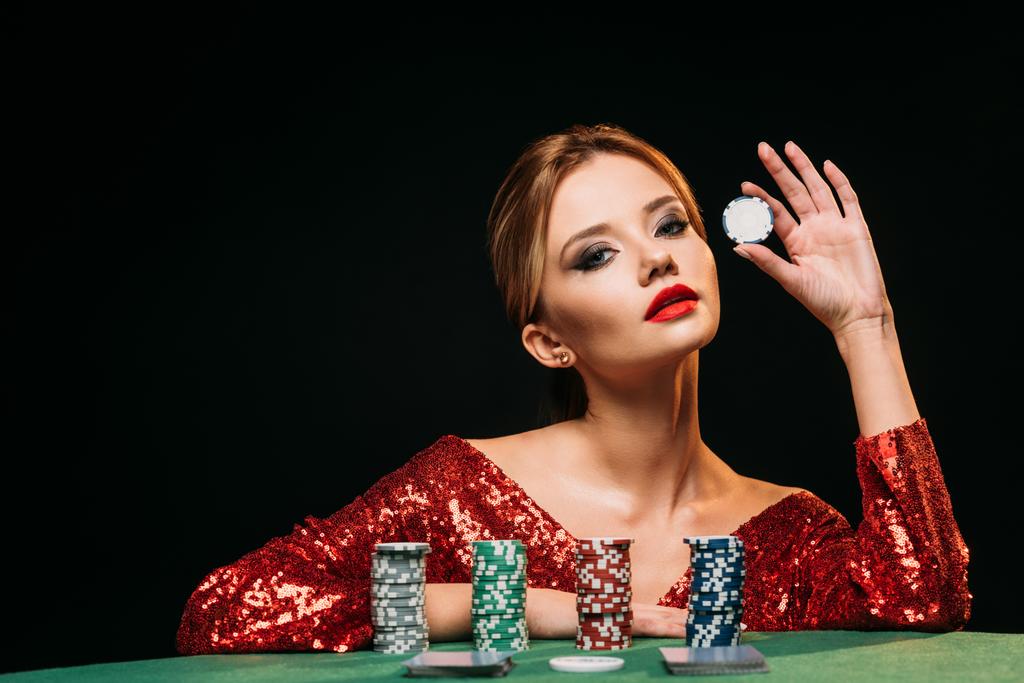 attractive girl in red shiny dress leaning on table, holding poker chip and looking at camera isolated on black - Photo, Image