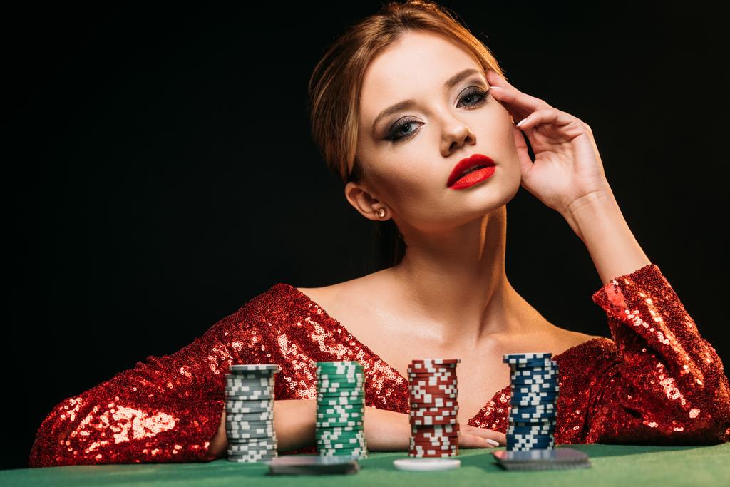όμορφο κορίτσι με κόκκινο λαμπερό φόρεμα ακουμπά στο τραπέζι με μάρκες πόκερ και να βλέπουν τα φωτογραφικών μηχανών που απομονώνονται σε μαύρο - Φωτογραφία, εικόνα
