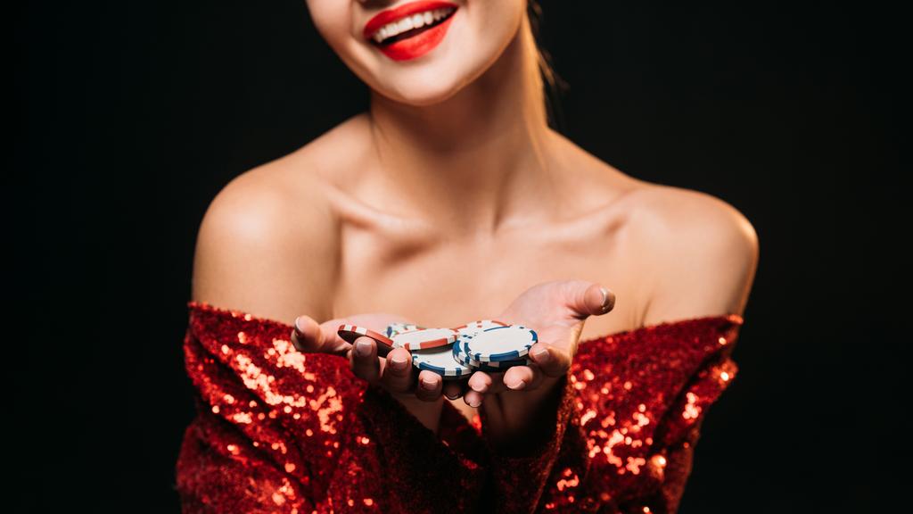 bijgesneden afbeelding van lachende meisje in de rode glanzende jurk bedrijf casinofiches geïsoleerd op zwart - Foto, afbeelding