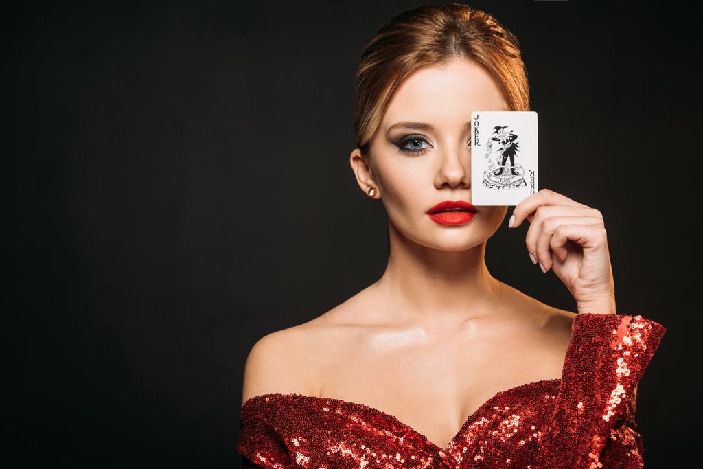 attractive girl in red shiny dress covering eye with joker card isolated on black - Photo, Image