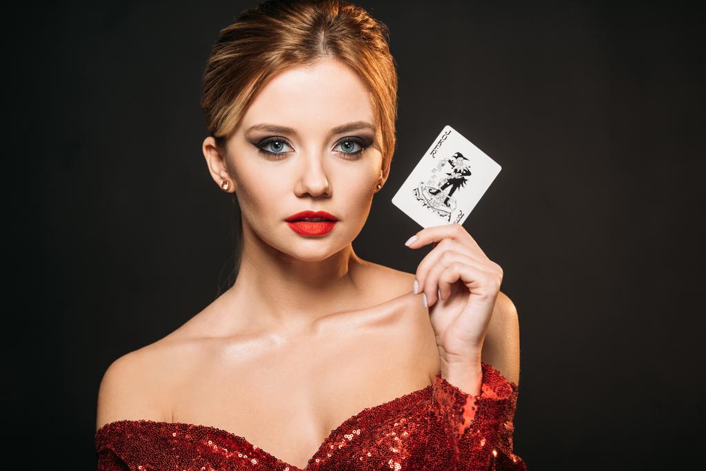 kırmızı parlak elbiseli joker kartı tutan ve siyahta izole kamera bakarak çekici kız - Fotoğraf, Görsel