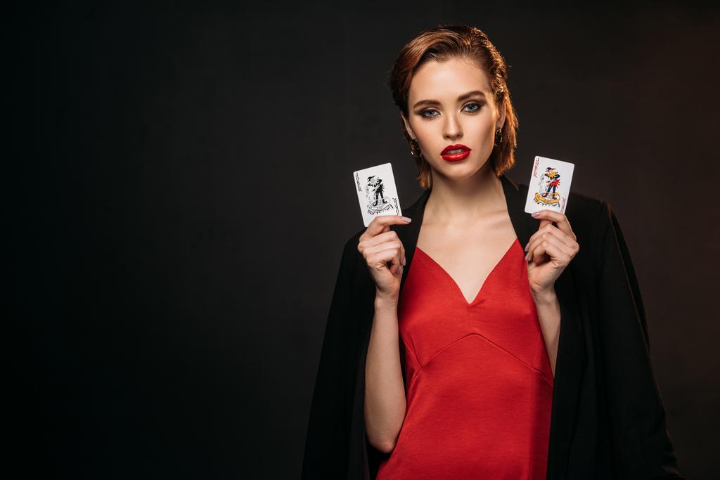 piękna dziewczyna w czerwonej sukience i czarna kurtka posiadania kart do pokera i patrząc na kamery na czarnym tle - Zdjęcie, obraz