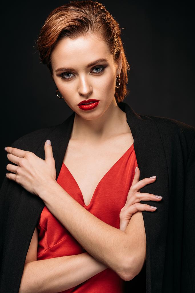 menina atraente em vestido vermelho e casaco preto de pé com braços cruzados e olhando para a câmera isolada no preto
 - Foto, Imagem