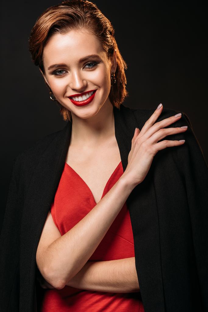 atractiva chica sonriente en vestido rojo y chaqueta negra mirando a la cámara aislada en negro
 - Foto, imagen