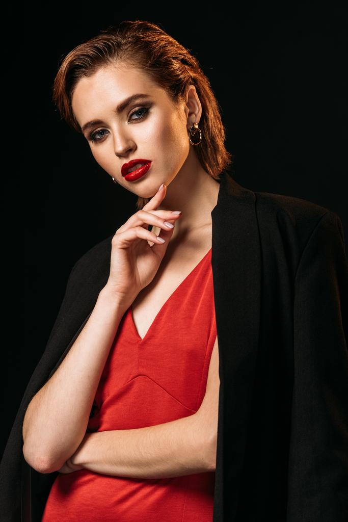 attractive girl in red dress and black jacket touching chin and looking at camera isolated on black - Photo, Image