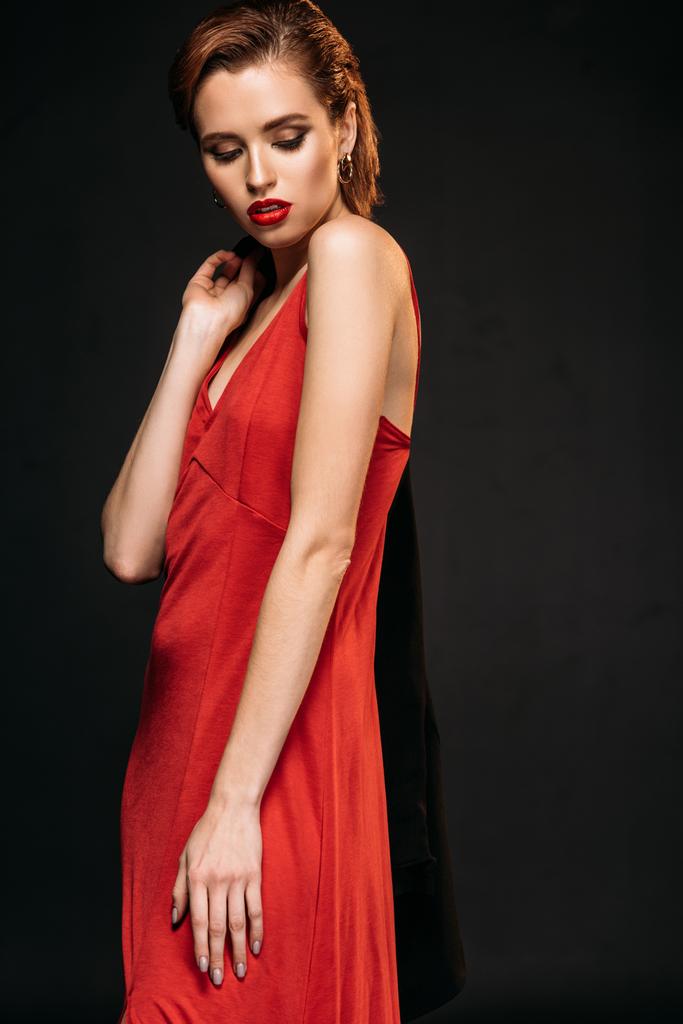 chica atractiva en vestido rojo sosteniendo chaqueta negra y mirando hacia abajo aislado en negro
 - Foto, imagen