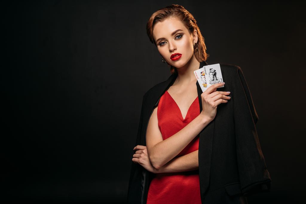 schönes Mädchen in rotem Kleid und schwarzer Jacke mit Spielkarten und Blick in die Kamera isoliert auf schwarz - Foto, Bild