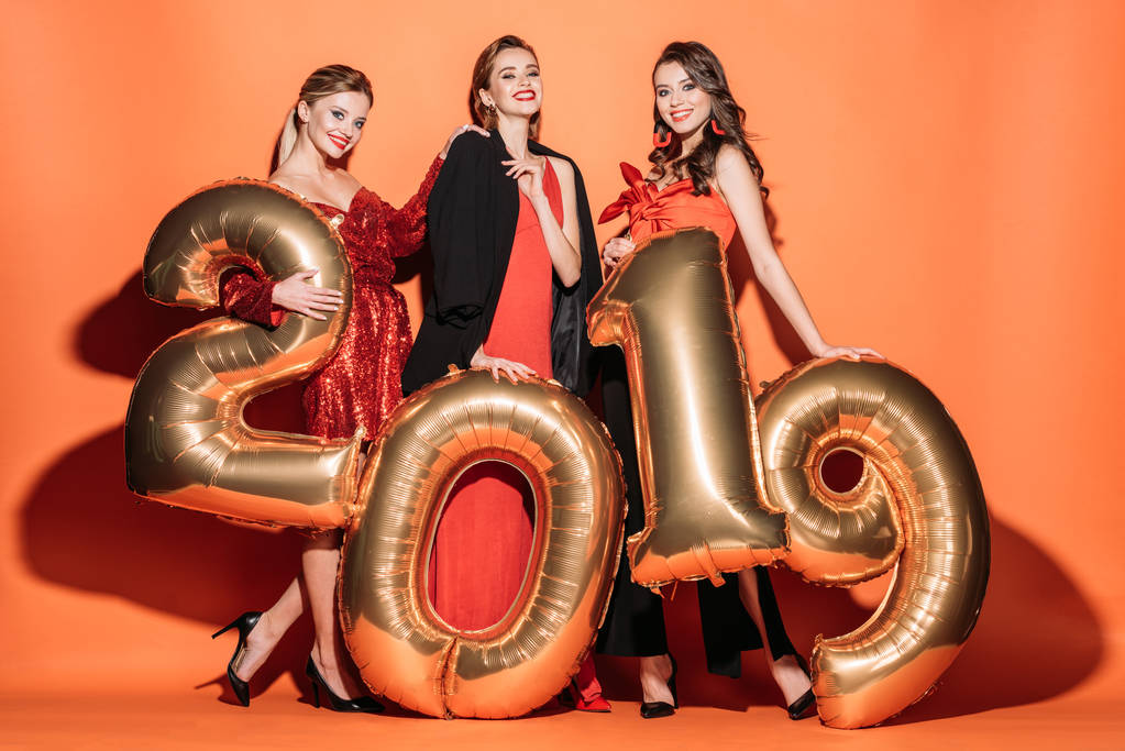 счастливые привлекательные девушки в стильной одежде для вечеринок, позирующие с золотыми шариками 2019 года на апельсине
 - Фото, изображение