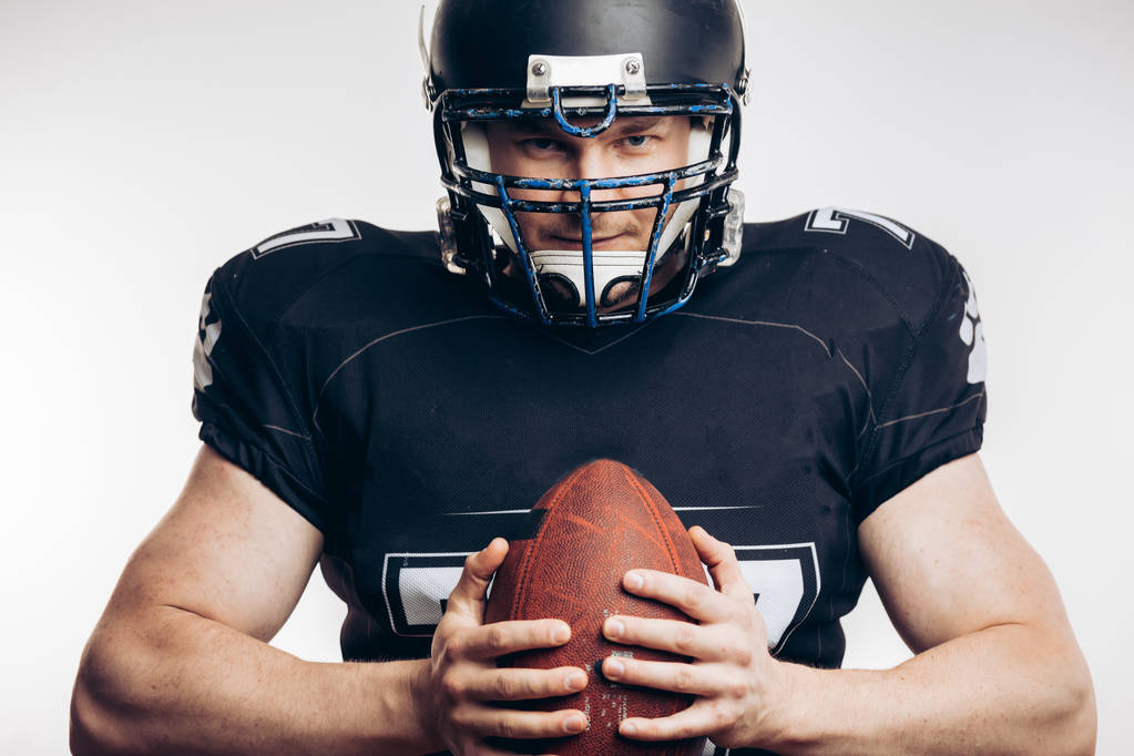 Мускулистый американский футболист в защитной форме и шлеме, держащий мяч - Фото, изображение
