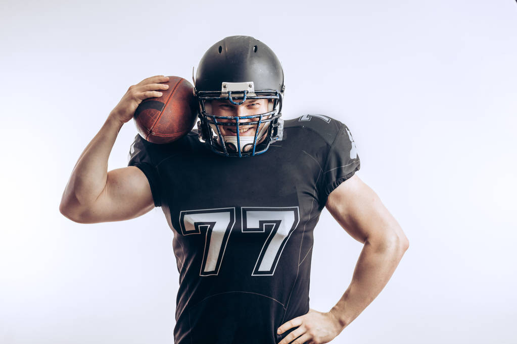 Giocatore di football americano muscolare in uniforme protettiva e casco con palla in mano - Foto, immagini