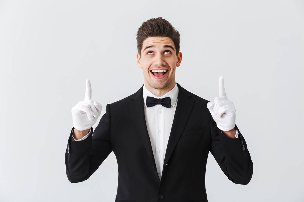 Portret przystojny młody mężczyzna kelner noszenie tuxedo i rękawice, stojąc na białym tle nad białym tle, wskazując palcami w miejsce - Zdjęcie, obraz