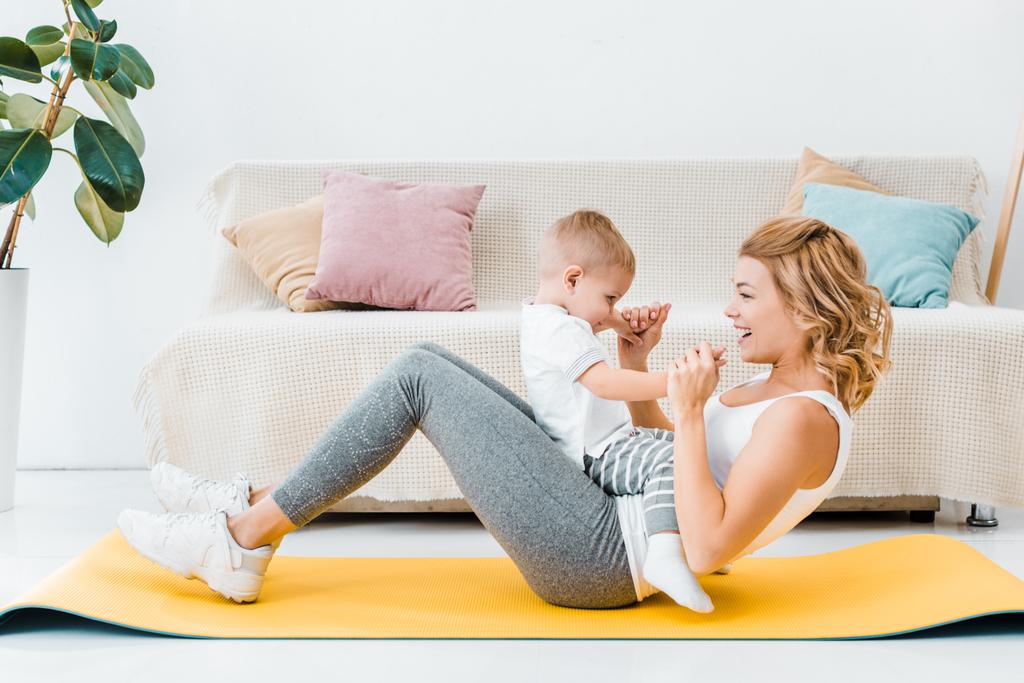 woman holding toddler son and training on fitness mat in living room - Photo, Image