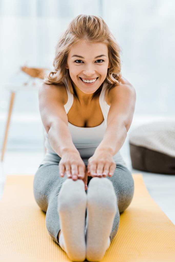 χαμογελαστό γυναίκα κάνει τεντώνοντας άσκηση σε γυμναστήριο Κίτρινο χαλί και κοιτάζοντας κάμερα στο σπίτι - Φωτογραφία, εικόνα