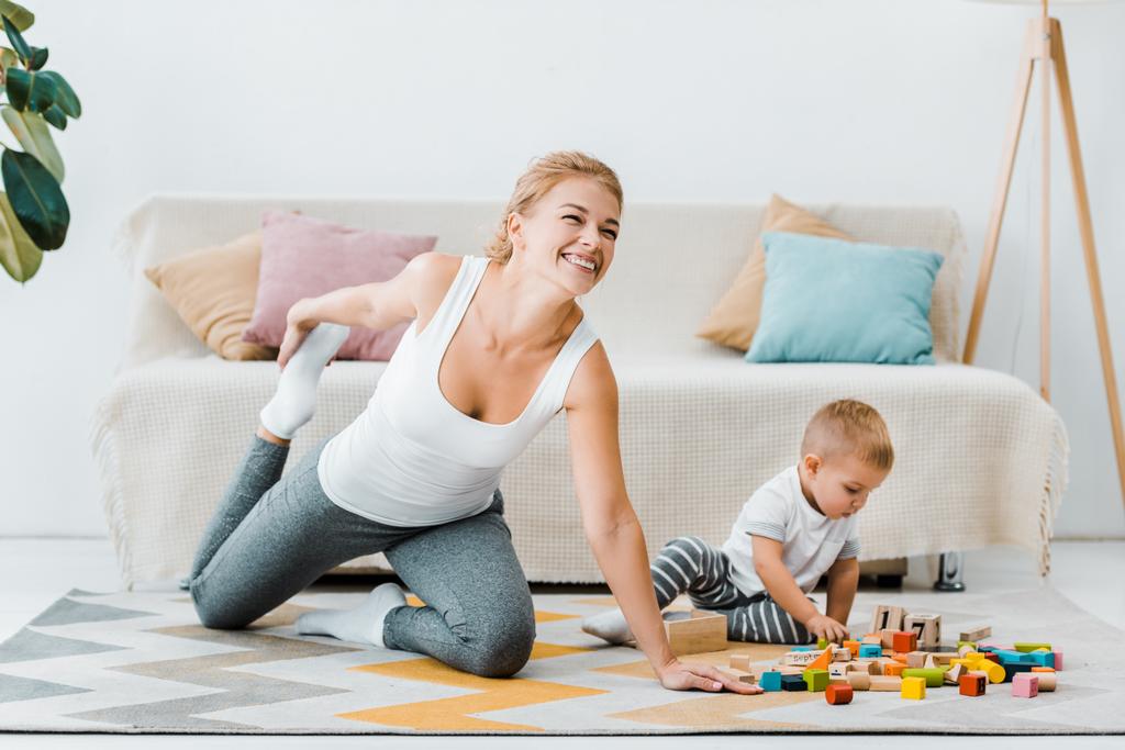 mãe sorridente alongamento perna e bonito filho criança brincando no tapete com cubos de madeira multicoloridos em casa
 - Foto, Imagem