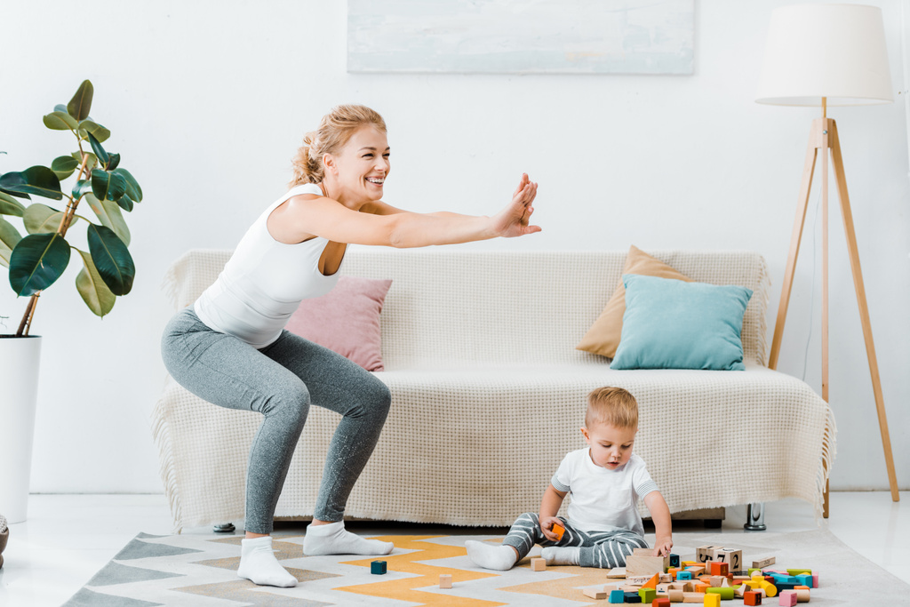 attractive mother doing squats and cute toddler son sitting on carpet and playing with multicolored cubes at home - Photo, Image